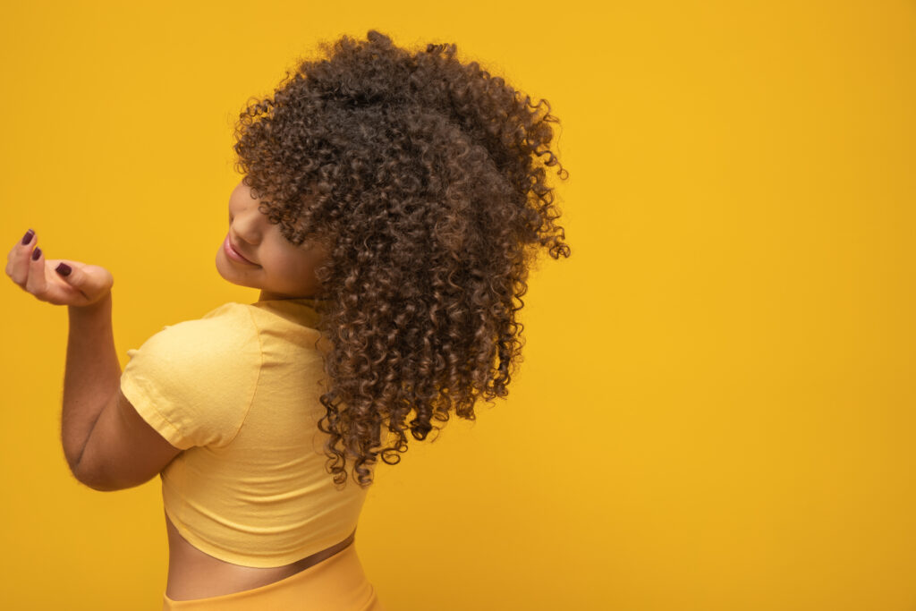 backwards american african woman with her curly hair on yellow background. laughing curly woman in sweater touching her hair and looking at the camera.