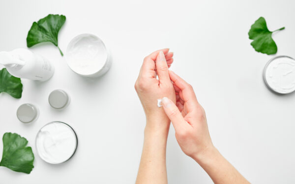 woman applying cosmetic moisturizing hand cream. cosmetic products, green leaves on white table. spa, manicure, skin care concept. flat lay, overhead view