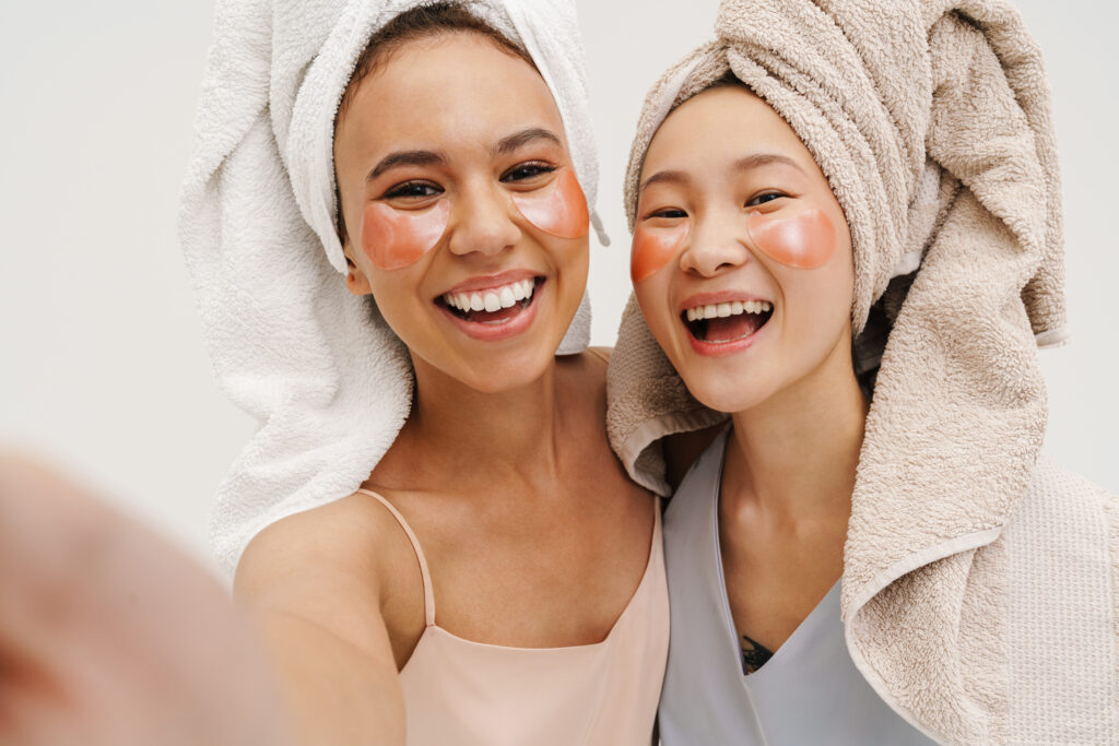 two cheerful women applying eye patches and taking selfie isolated over white wall