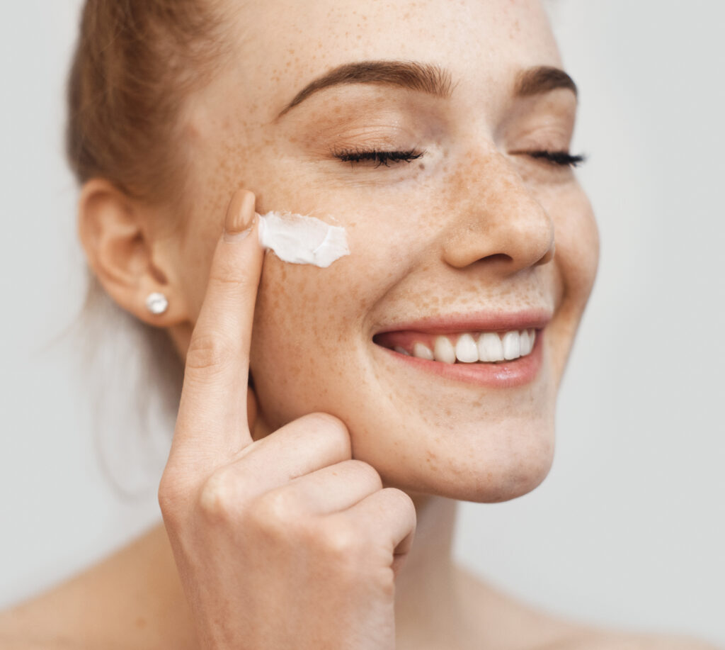 lovely caucasian woman with red hair and freckles applying anti aging cream on her face while posing on a beige wall