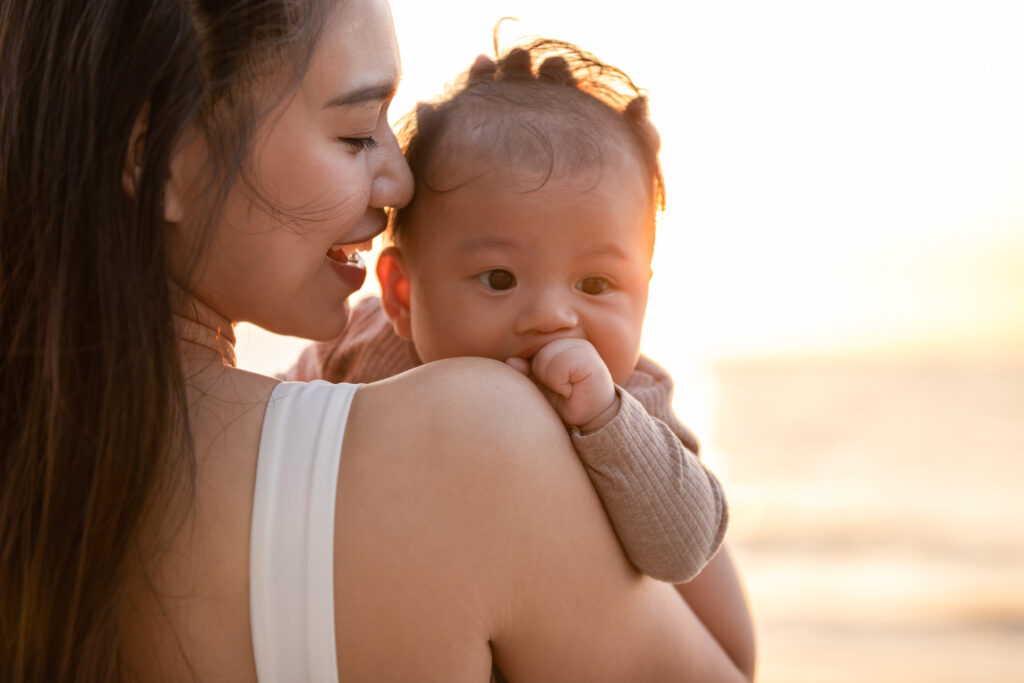 beautiful attractive asian mom holding her baby newborn in hand and kissing on baby head sweet and lovely outdoor on the beach.happy mother and infant baby looking together smile with natural sunset